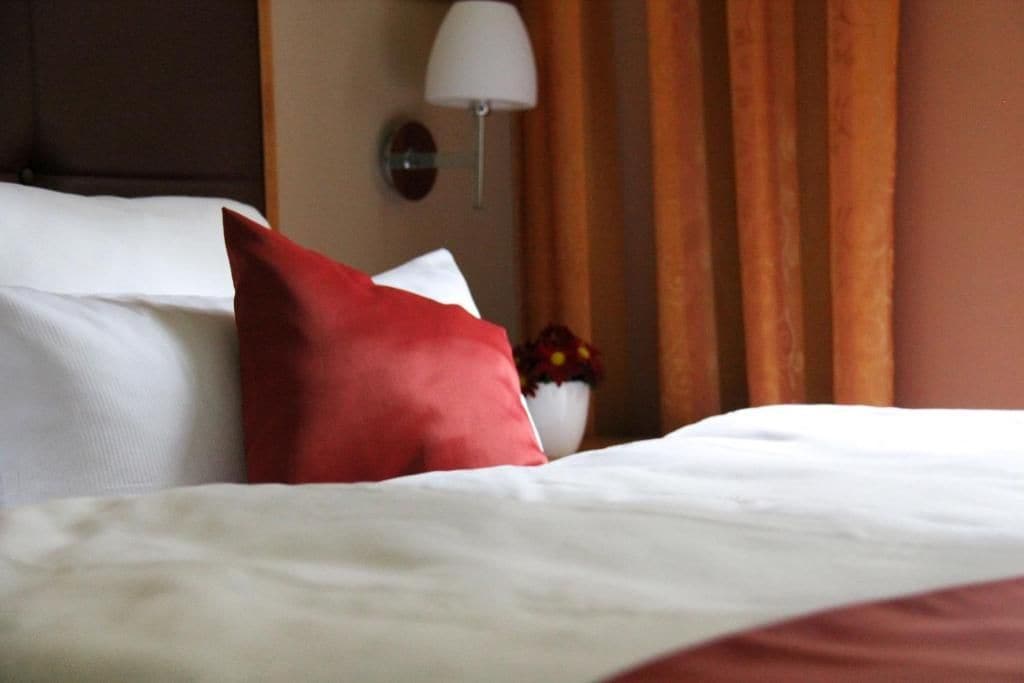 A hotel room with a neatly made bed, a red pillow, a white lamp, and a vase with flowers.