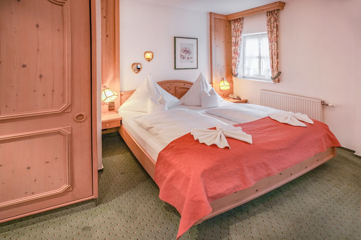 Una cama bien hecha con una colcha roja y almohadas blancas, un cabecero de madera, una mesita de noche con una lámpara, un cuadro en la pared y una ventana con cortinas.