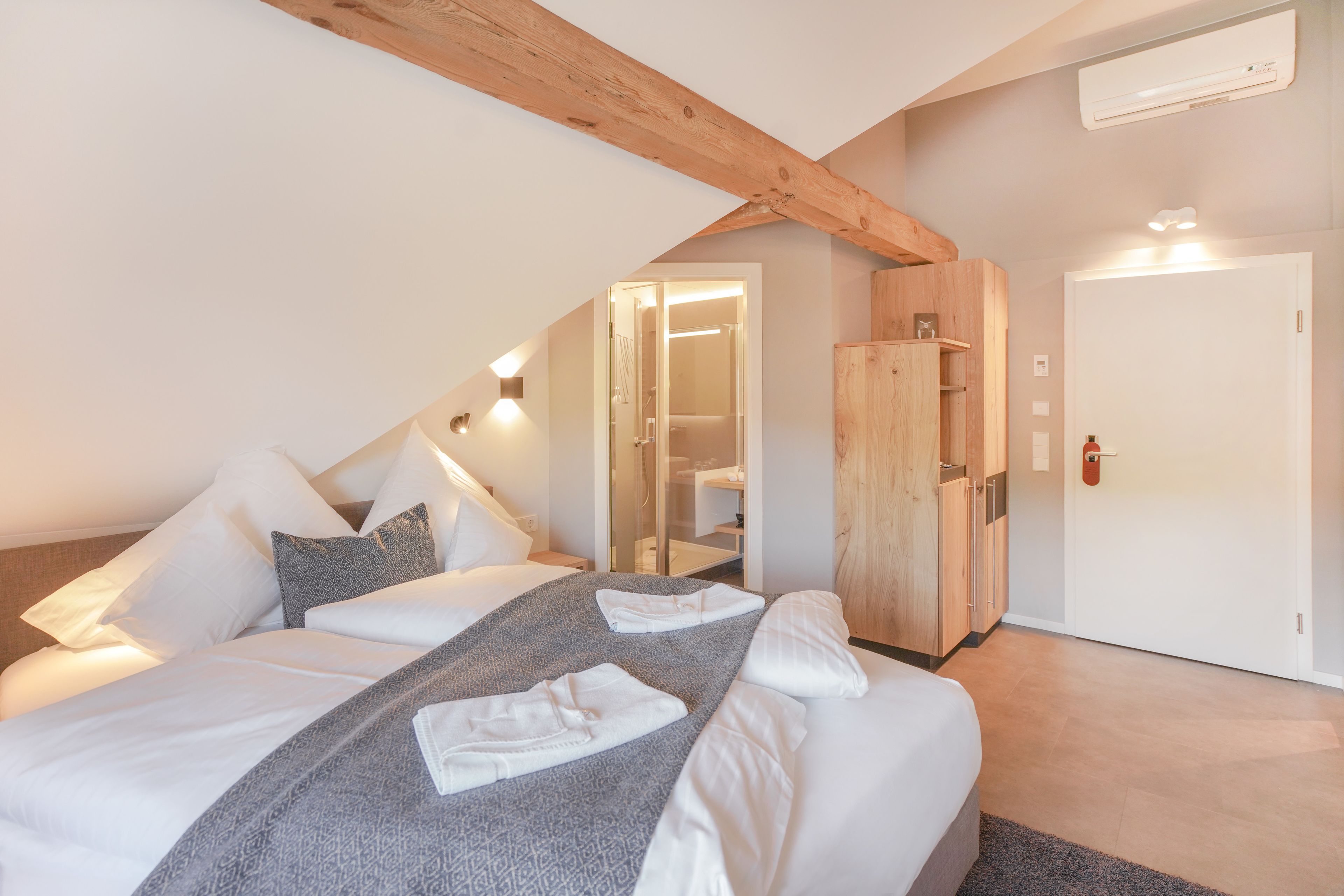 Ein Schlafzimmer mit einem großen Bett, einem Holzbalken an der Decke, einem Kleiderschrank und einem durch eine offene Tür einsehbaren Badezimmer.
