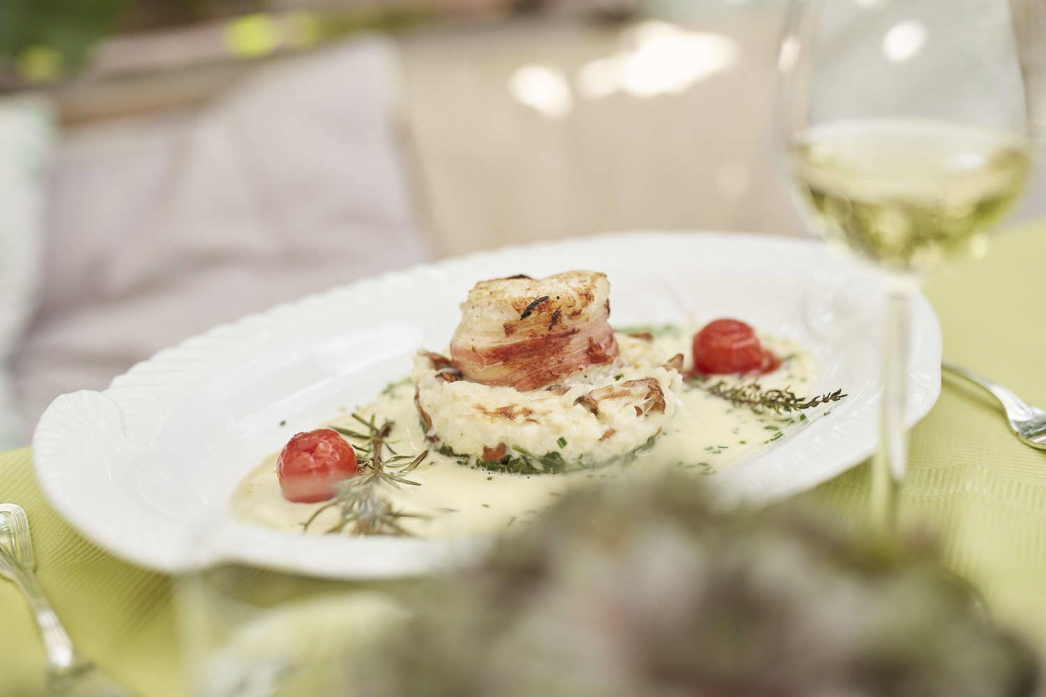 Un plato blanco con un guiso, un tenedor y una copa de vino blanco sobre una mesa.