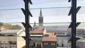 A view through a window with a metal frame, featuring a variety of buildings including a church with a spire and several residential buildings with different roof styles. - LV Premier Marques - MQ2 (Porto)