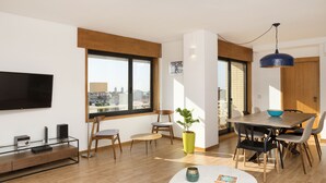 A modern living room with a flat-screen TV, wooden flooring, a dining area with a table and chairs, a potted plant, and large windows offering a city view. - LV Premier Marques - MQ2 (Porto)