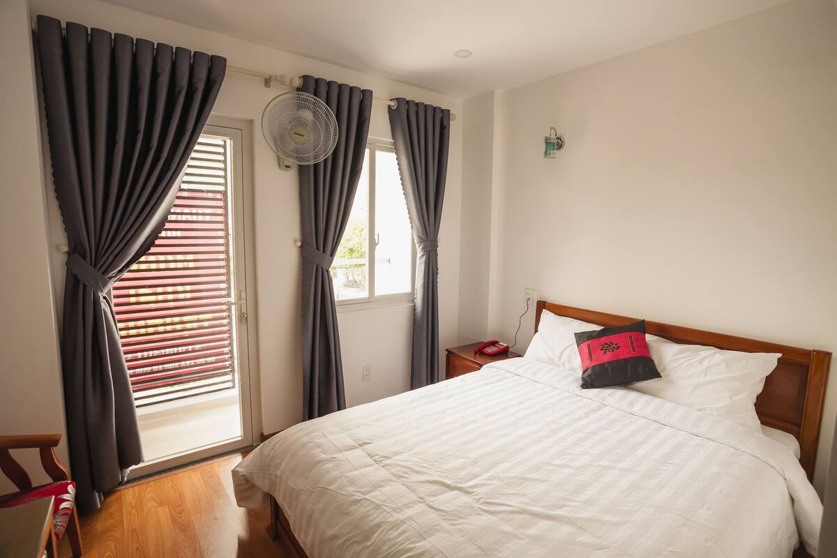 A bedroom with a bed, a nightstand, a fan, and a window with curtains.
