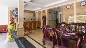 Um saguão de hotel com uma mesa de recepção, cadeiras de madeira, um relógio e um arranjo floral.