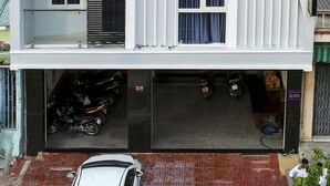 Um hotel moderno com uma placa de destaque que diz "THANH THANH HOTEL". Possui várias varandas com plantas em vasos, uma área de estacionamento coberta e um carro branco estacionado na frente.