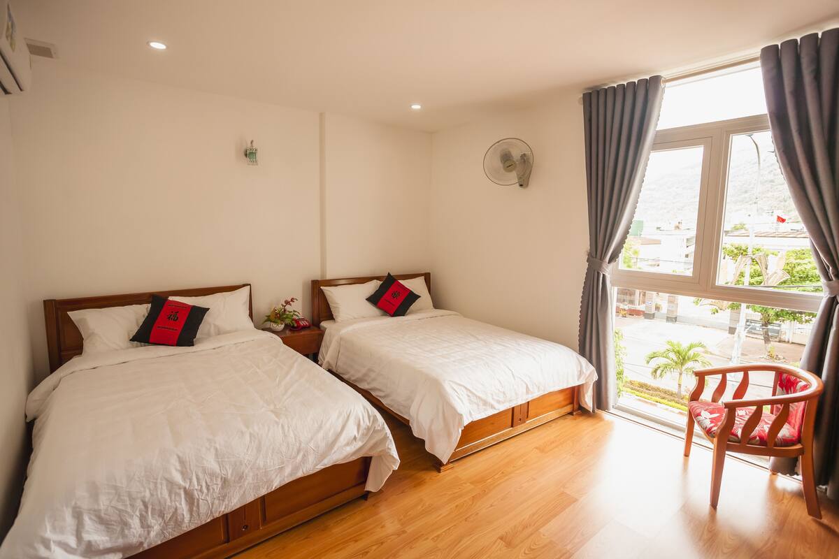 A hotel room with two beds, a wooden floor, a window with curtains, and a chair.