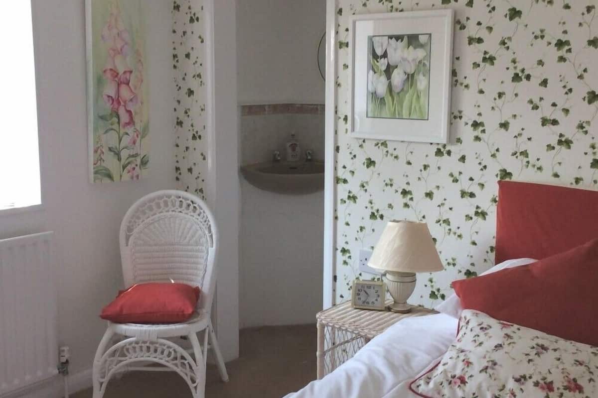 A bedroom with a bed, a chair, a nightstand with a lamp, and a wall with floral wallpaper.