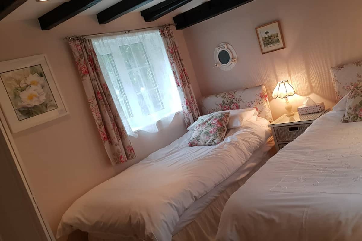 A bedroom with two beds, floral bedding, a window with curtains, a bedside lamp, and a framed picture on the wall.