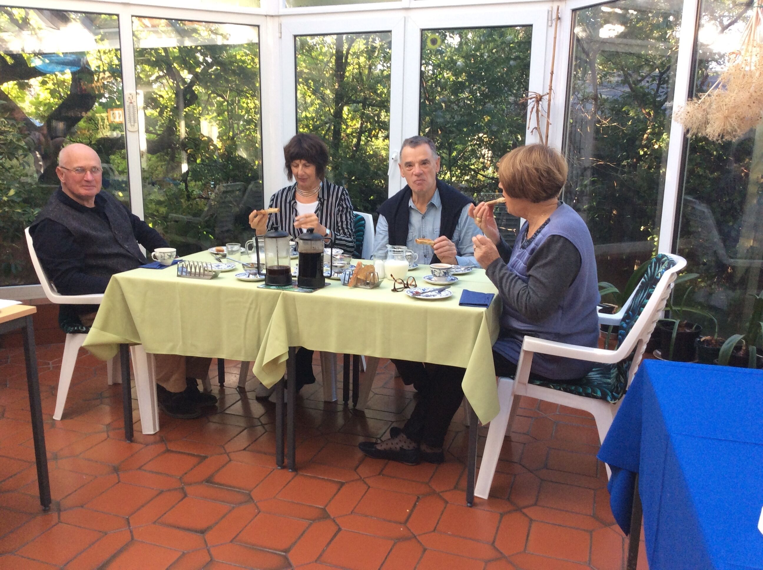 A group of people dining at an outdoor table with a green tablecloth, surrounded by glass walls and potted plants.