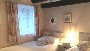 Une chambre avec deux lits simples, des rideaux à motifs floraux, une fenêtre ronde et une table de chevet avec une lampe.