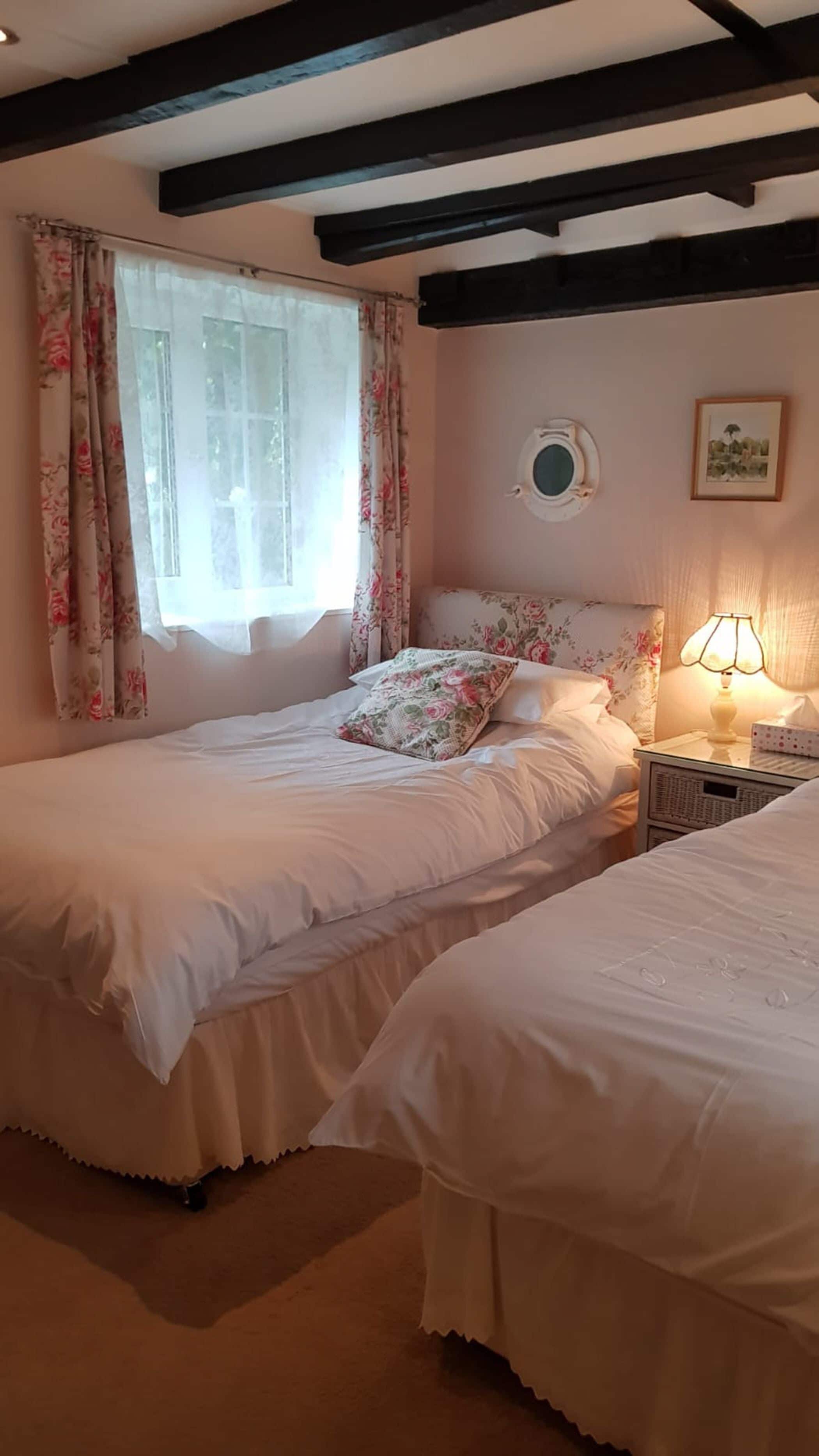 Ein Schlafzimmer mit zwei Betten, blumigen Vorhängen, einem Rundfenster, einer Nachttischlampe und einem gerahmten Bild an der Wand.