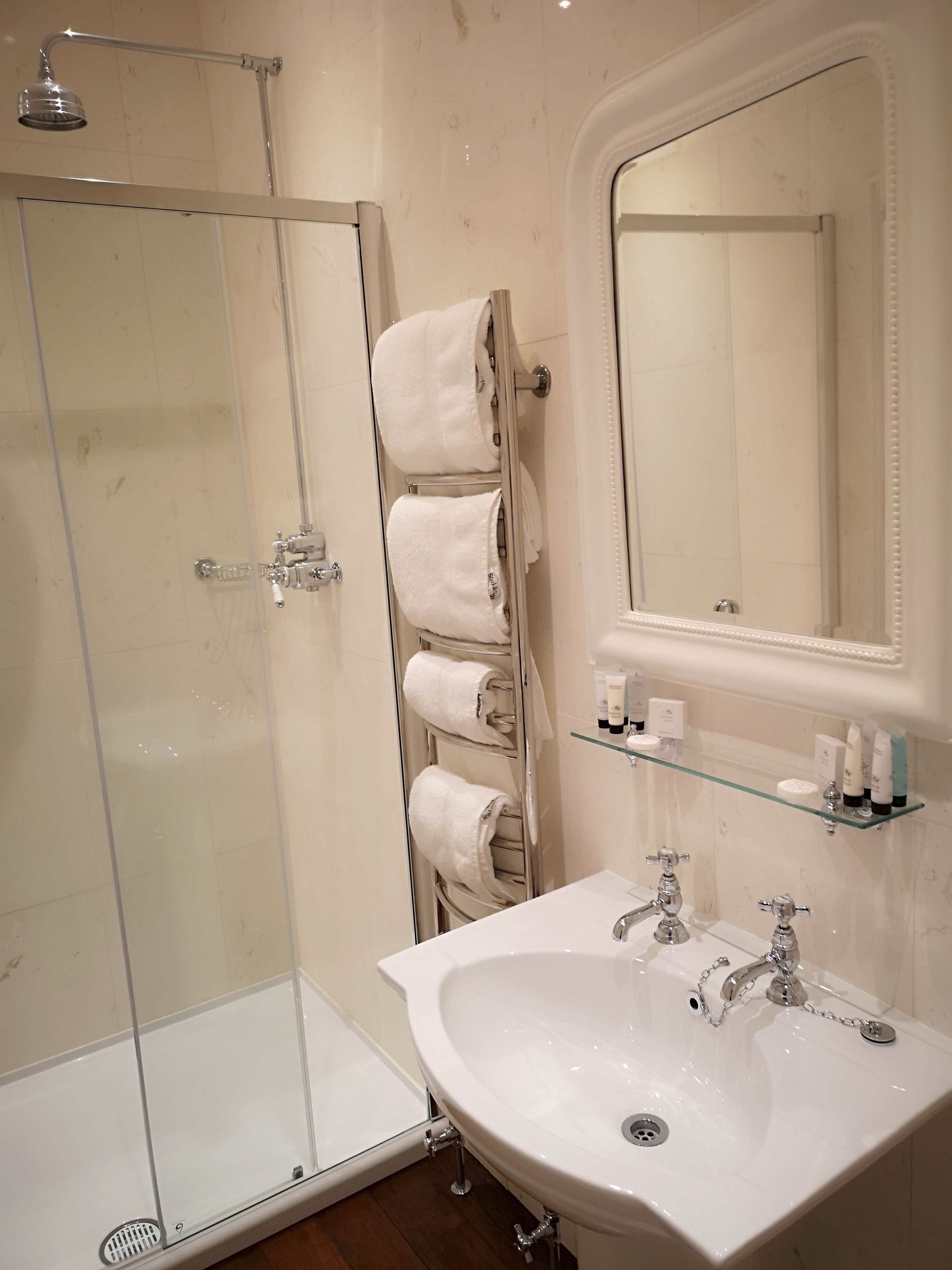Un baño con cabina de ducha de vidrio, un lavabo blanco con grifería cromada y un perchero con varias toallas blancas.