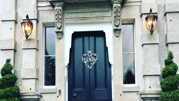 A dark blue door with a heart-shaped decoration and the word "LOVE" in the center, flanked by two wall-mounted lamps and two potted plants. - Cleatham Hall (Gainsborough)