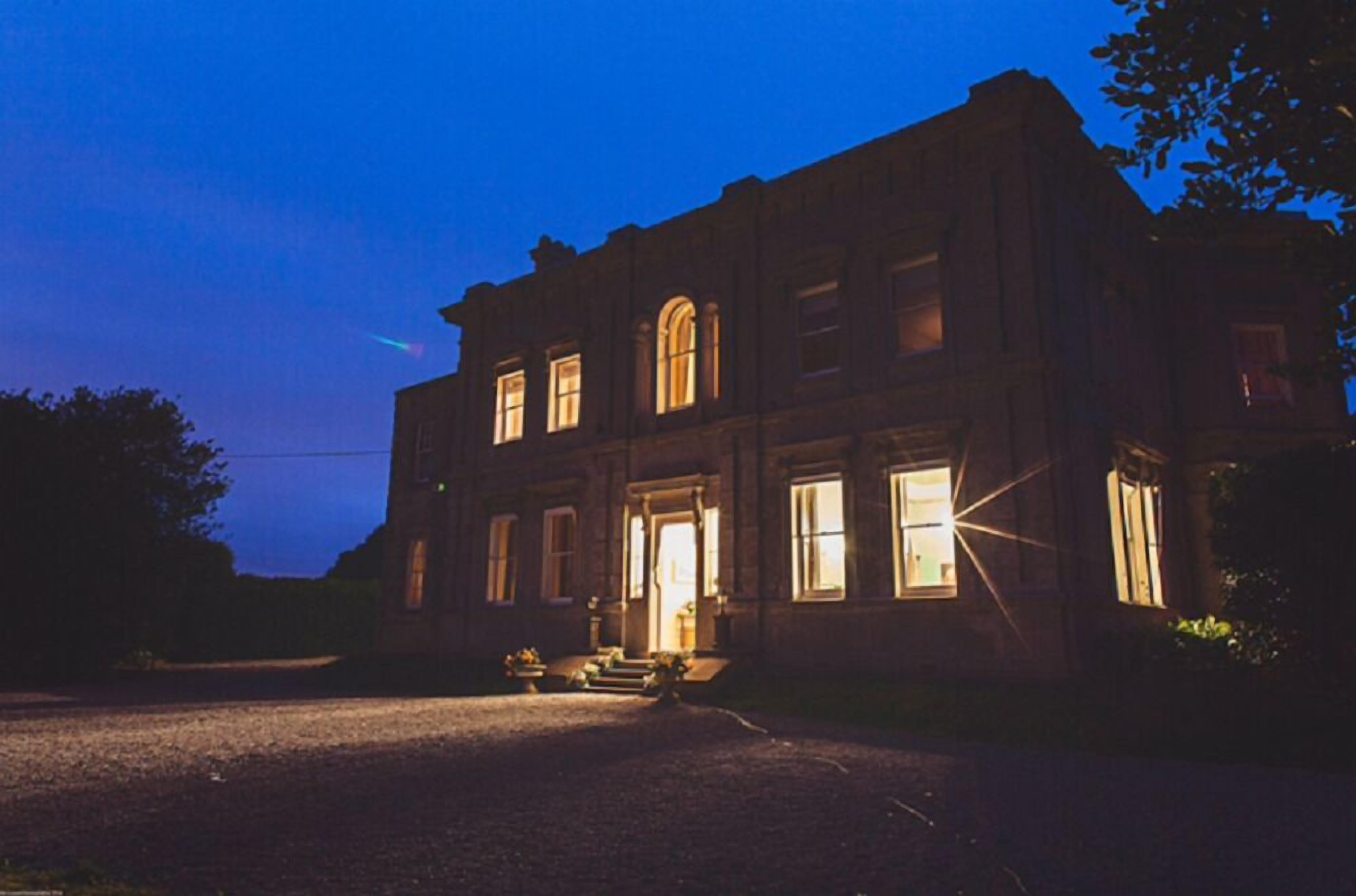 A large, historic building with a prominent entrance and multiple windows, illuminated by warm interior lighting.