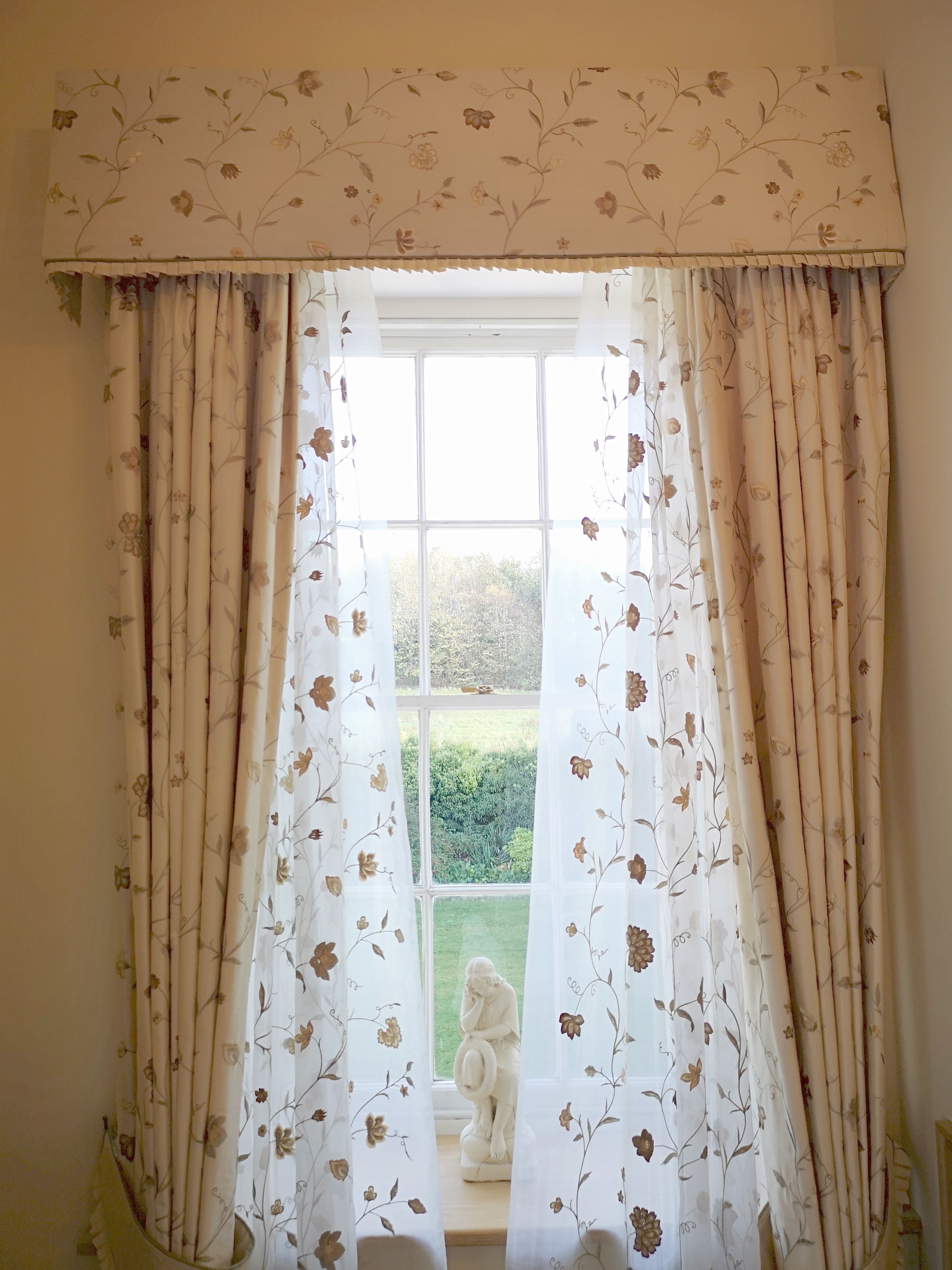 A room with a window covered by floral curtains, a white statue on the windowsill, and a view of a green garden.