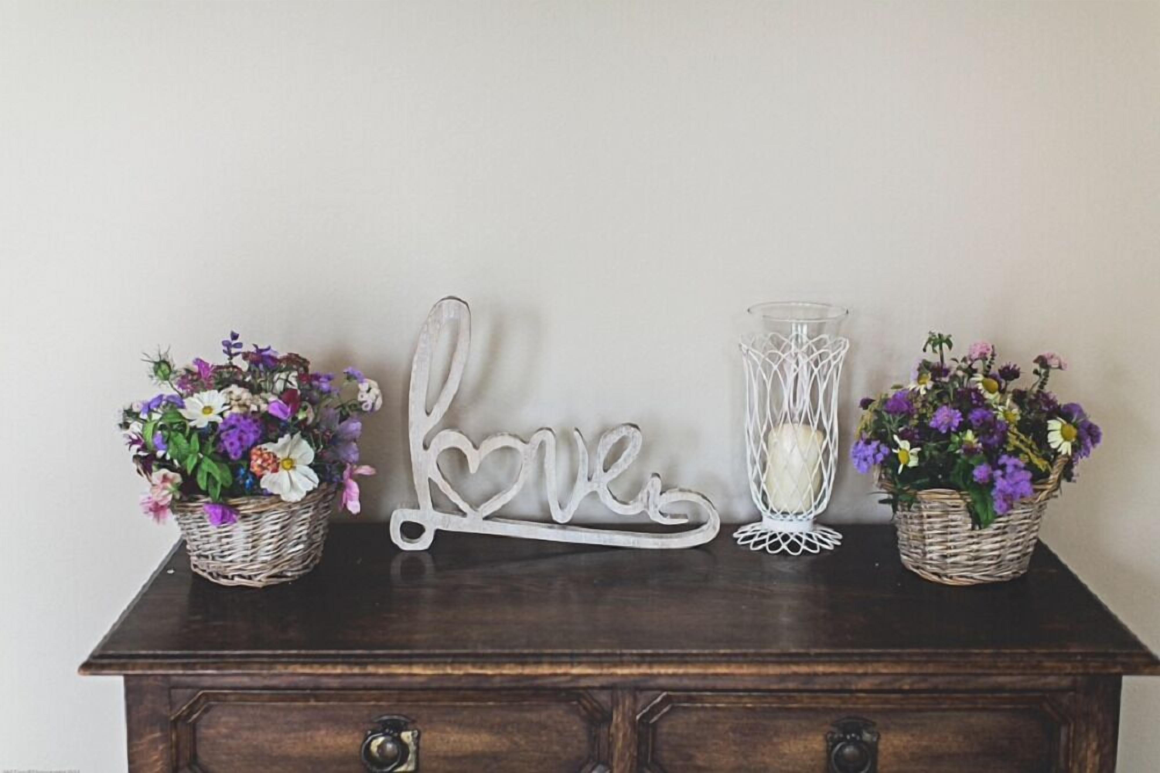 Un aparador de madera oscura con dos cestas de mimbre que contienen flores moradas y blancas, un cartel blanco con la palabra 'Love', y un florero transparente de base blanca.