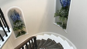 A staircase with ornate railings and a white wall with two arched windows, each containing a floral arrangement. - Cleatham Hall (Gainsborough)
