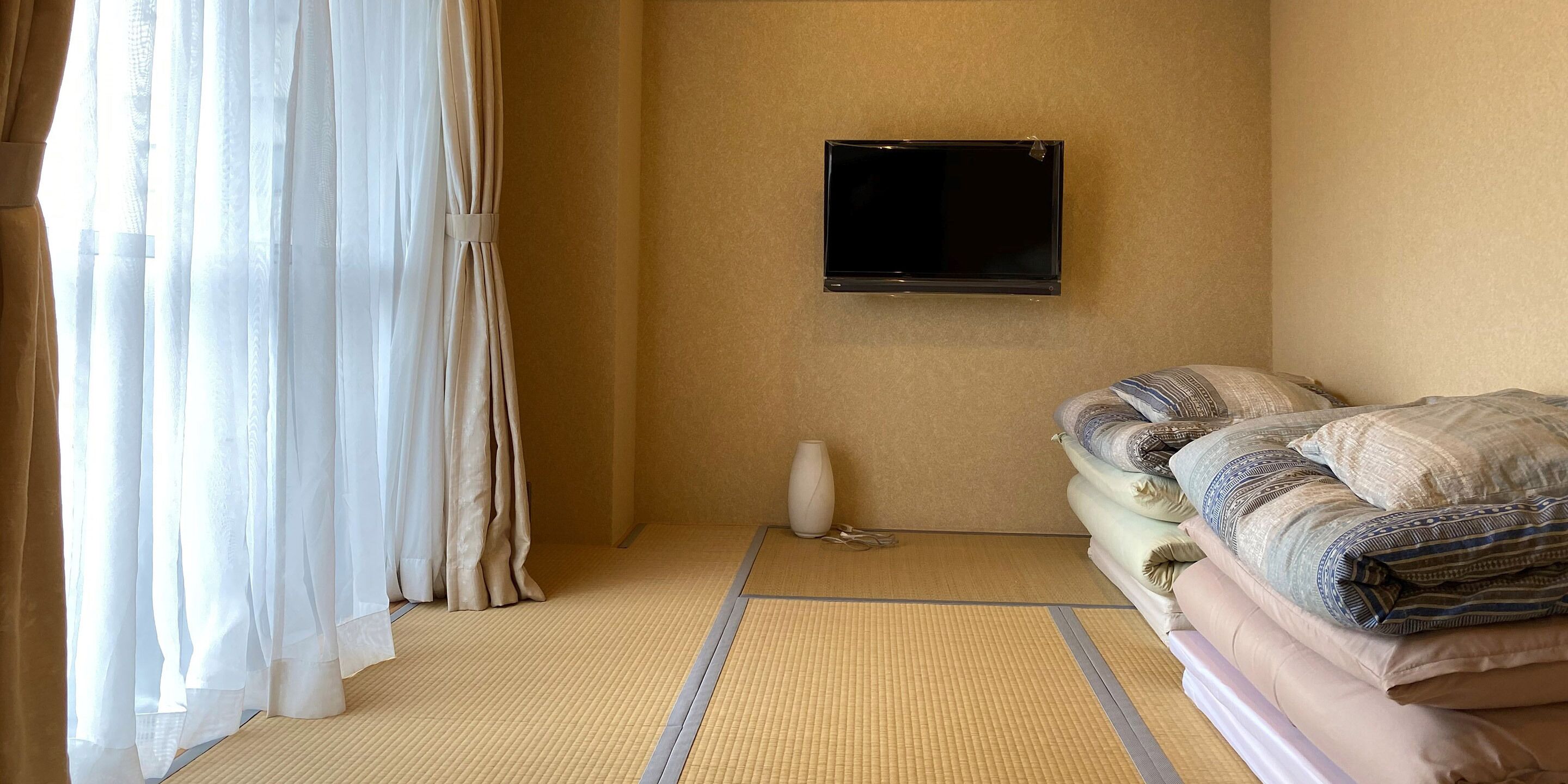 A hotel room with tatami flooring, a bed with patterned bedding, a mounted flat-screen TV, and a vase on the wall.