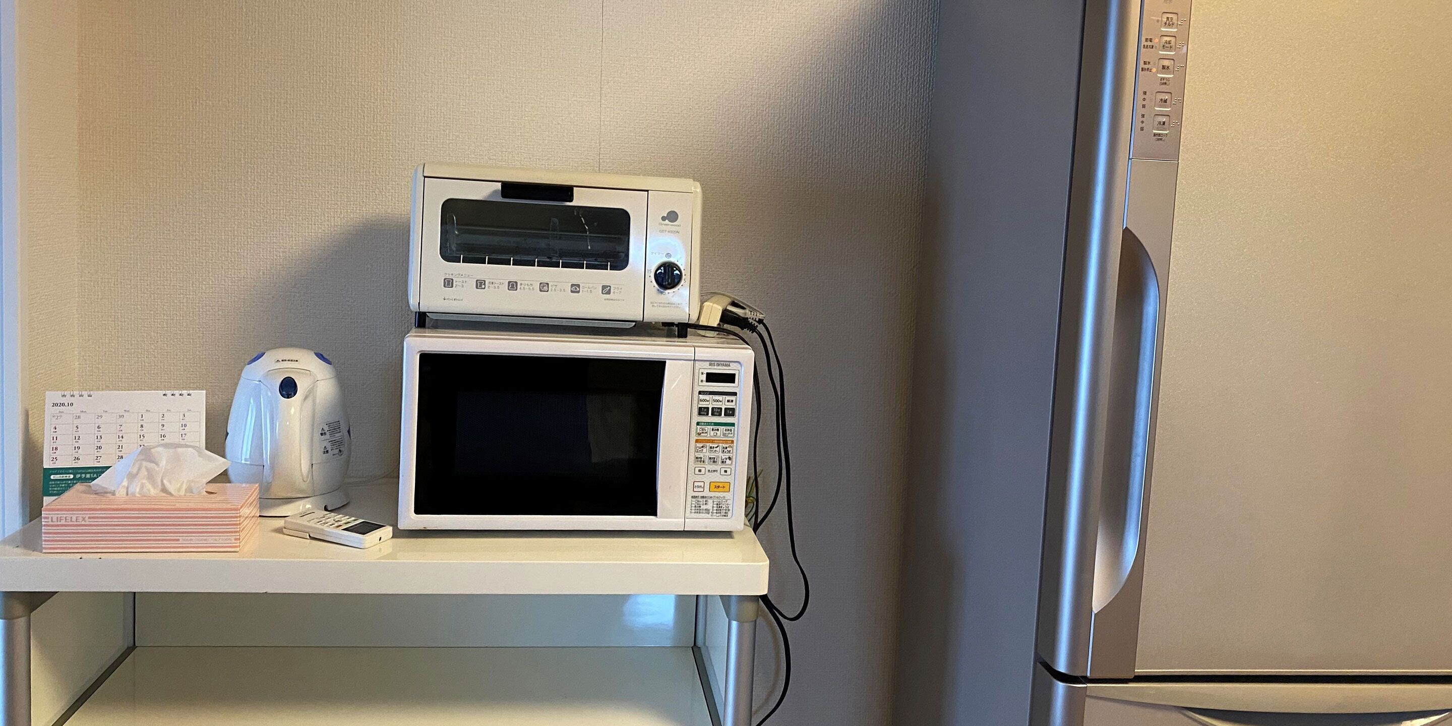 Un four à micro-ondes doté d’un panneau de commande et d’un écran numérique, un calendrier, une boîte à mouchoirs et une bouilloire, le tout posé sur un plan de travail blanc.