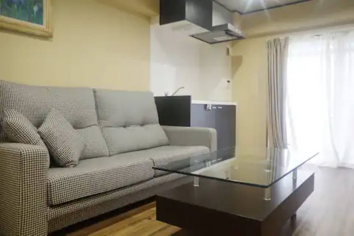 Ein Wohnzimmer mit einem karierten Sofa, einem Glastisch, einem dunklen Holzboden, einer Küchenzeile mit Schränken und einem Fenster mit Vorhängen.