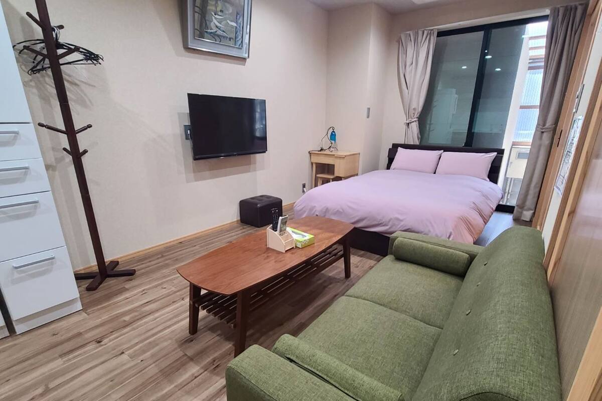 Ein modernes Hotelzimmer mit einem grünen Sofa, einem Holztisch, einem Flachbildfernseher, einem Bett mit rosa Bettwäsche, Holzboden und einem Kleiderhaken.