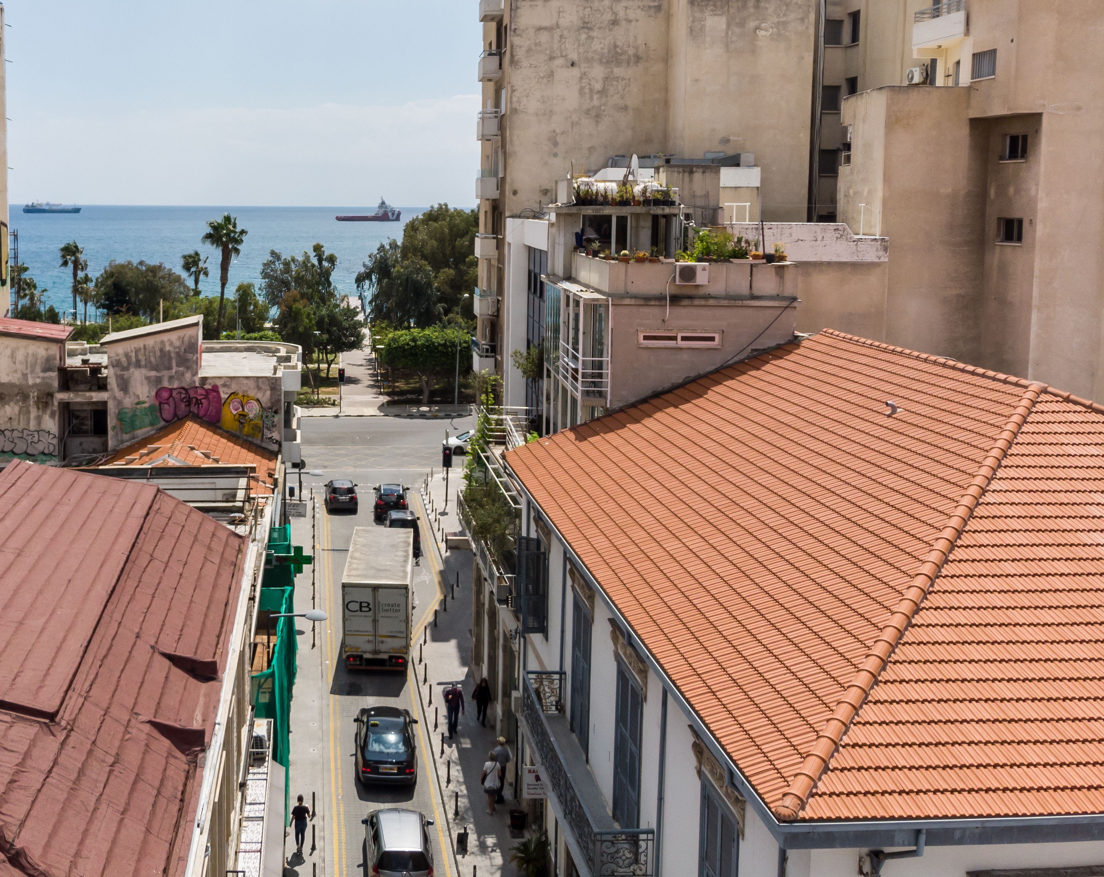 A street view with a building featuring a red-tiled roof, a visible balcony, and a clear view of the sea with a ship in the distance.