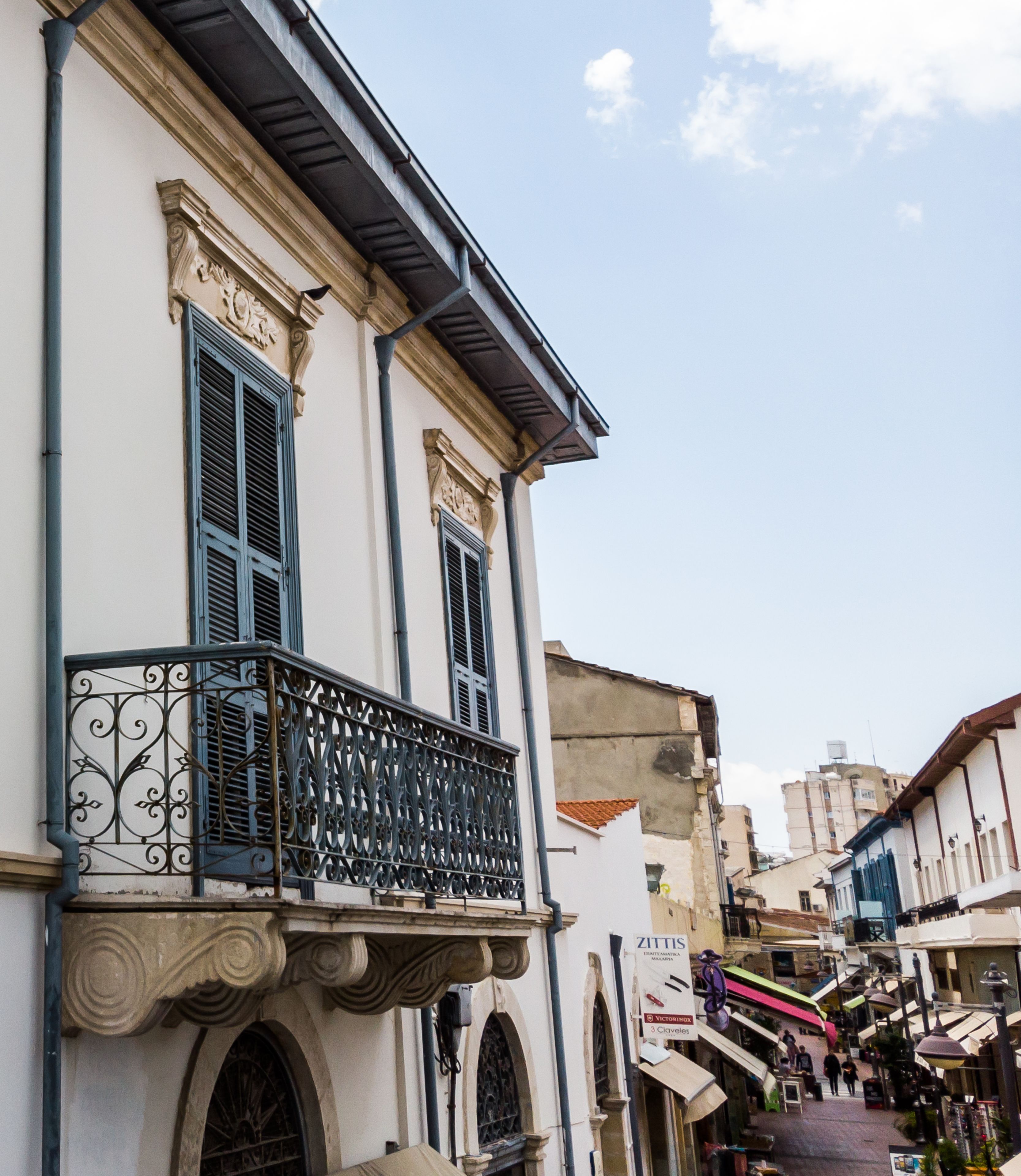 A narrow street with a building featuring a balcony with ornate metal railings and a shop with a sign.