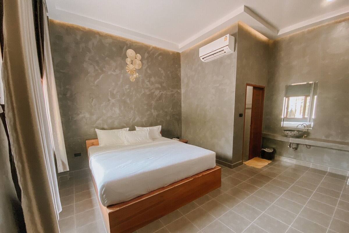 Quarto moderno com cama de madeira, ar condicionado integrado e banheiro com pia e espelho.