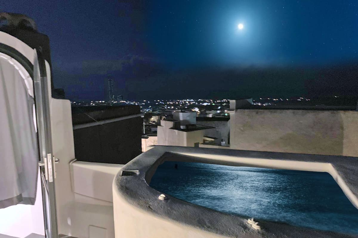 Un área de piscina en la azotea con vistas a la ciudad de noche y una luna llena en el cielo.