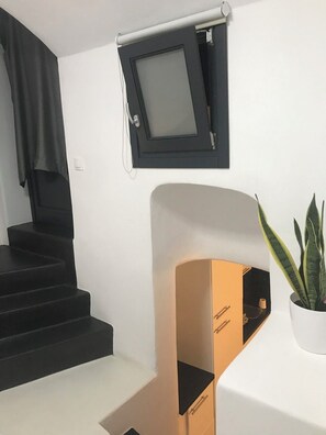 A staircase with black steps leading to a landing, a white wall with a black-framed window, and a potted plant on a white surface. - The Luna Suites (Santorini)