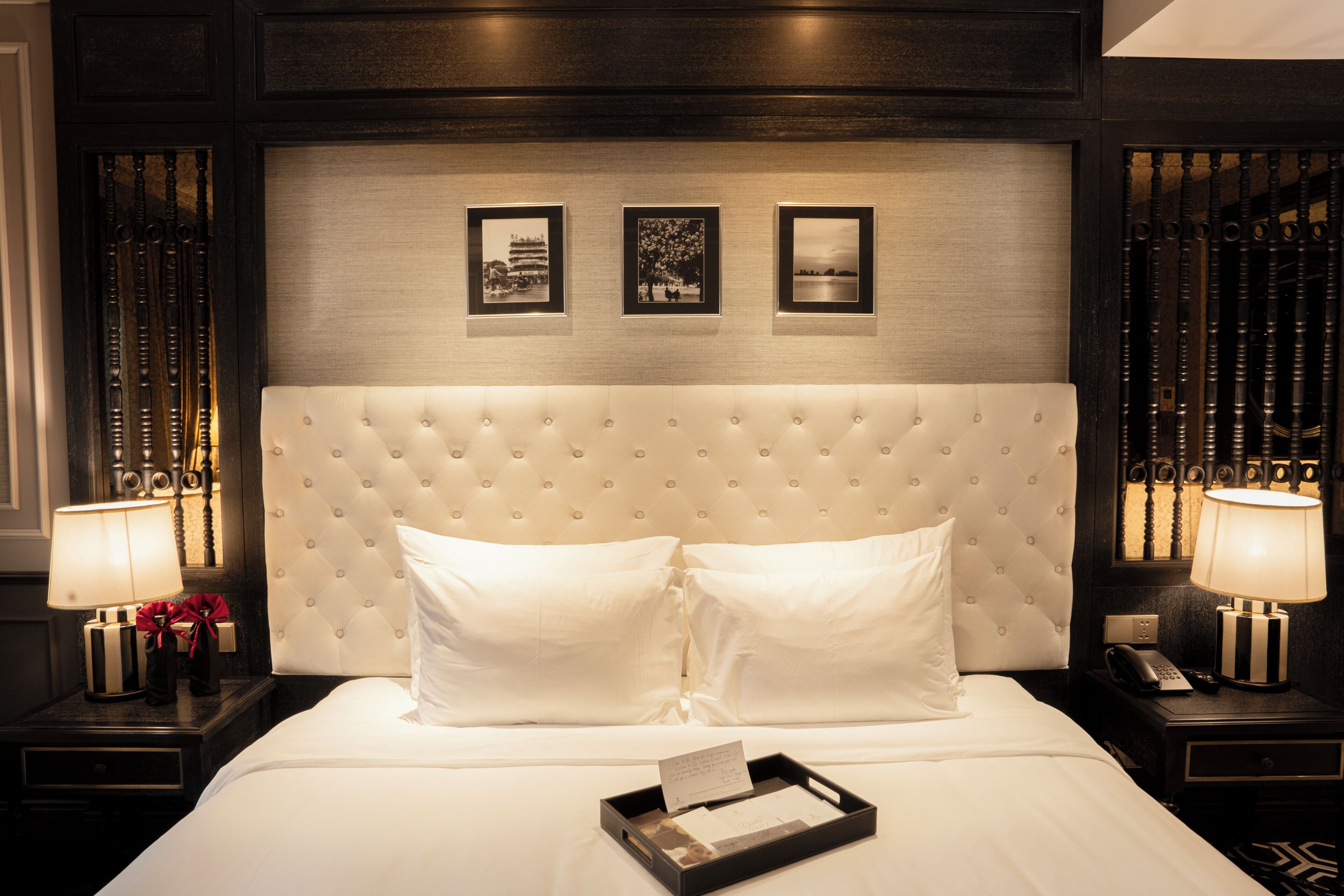Habitación de hotel con cabecera capitonada, ropa de cama blanca, dos lámparas de noche y cuadros en la pared.