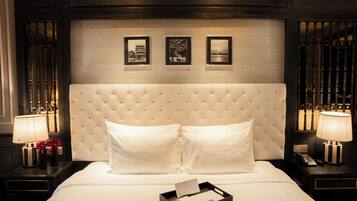 Habitación de hotel con cabecera capitonada, ropa de cama blanca, dos lámparas de noche y cuadros en la pared.