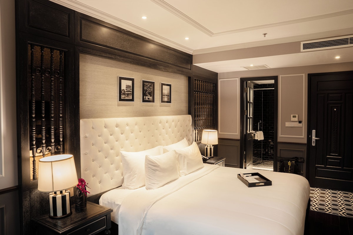 Quarto de hotel com uma cama grande, mesa de cabeceira com abajur, espelho e porta que dá para outro aposento.