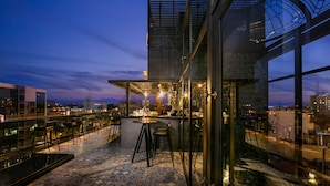 Un bar situé sur le toit d’un immeuble, avec une terrasse et une vue sur les toits de la ville au crépuscule.