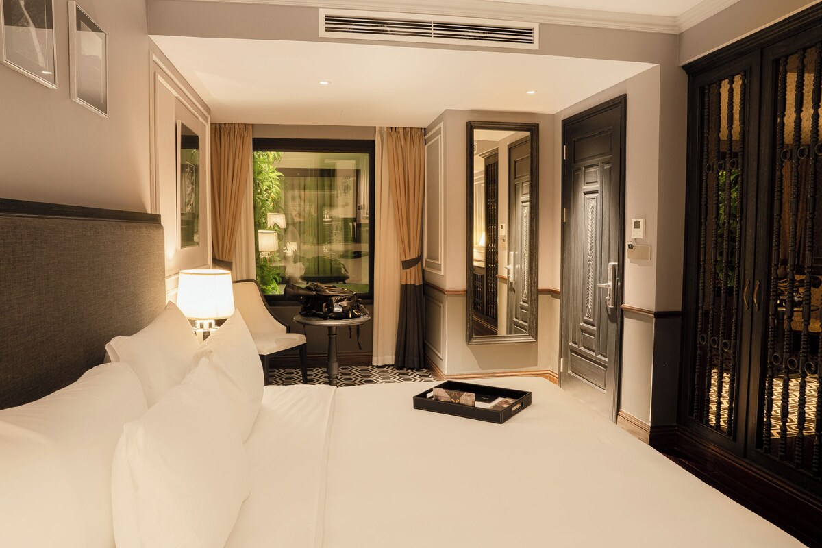 Quarto de hotel com uma cama, uma mesa de cabeceira com abajur, uma pequena mesa com uma planta e uma porta que dá para outro quarto.