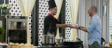 A chef serving food to a guest in a modern kitchen with a decorative window and a dining area with a table set for two.