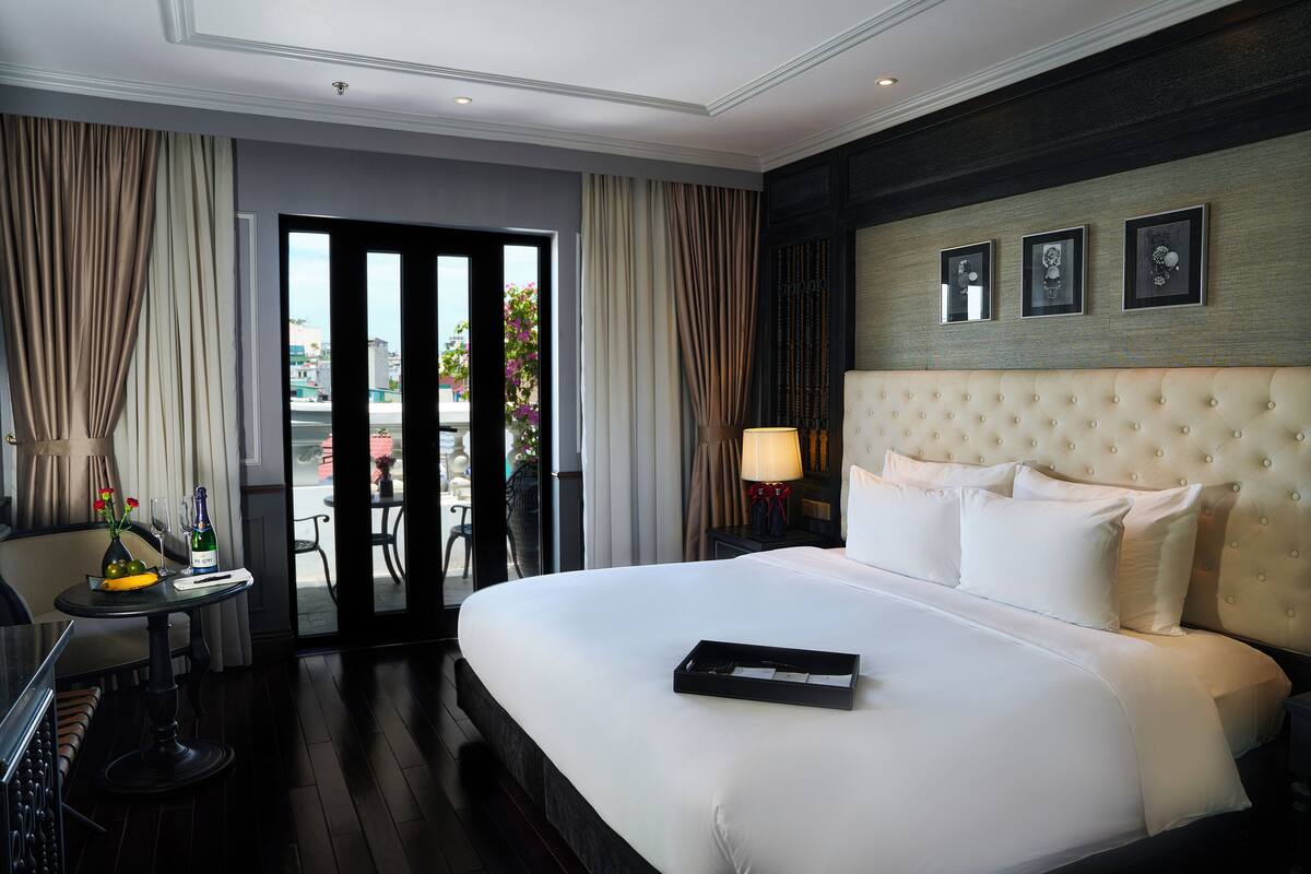 Ein Hotelzimmer mit einem großen Bett, einem kleinen Tisch mit Flasche und Gläsern sowie Blick durch eine Glastür nach draußen.