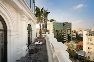 A balcony with a table and chairs, a palm tree, and a view of a modern building and surrounding cityscape.