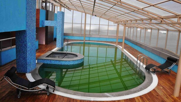 An indoor swimming pool with a glass ceiling, wooden flooring, and lounge chairs.