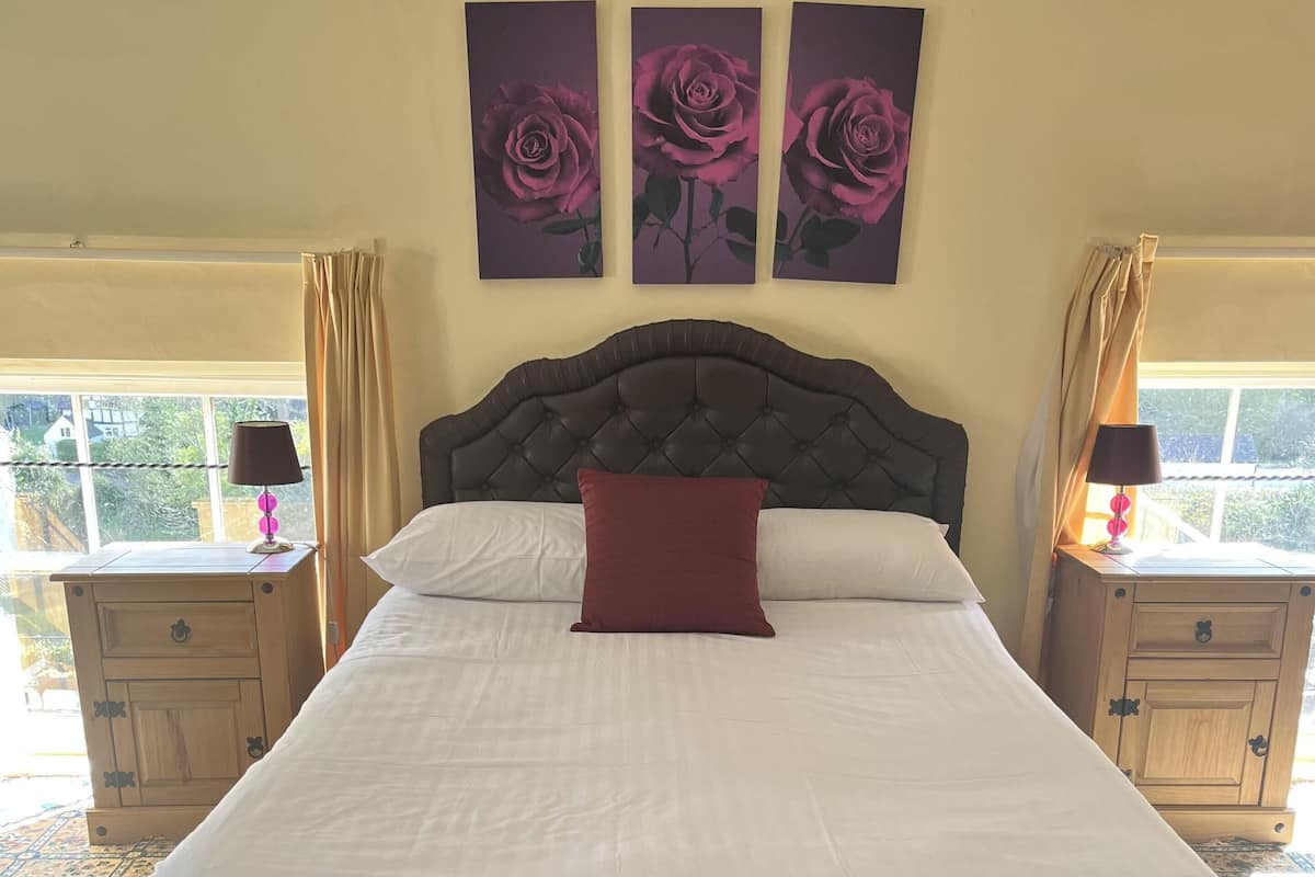 A large bed with a dark headboard, white linens, and a red accent pillow. There are two bedside tables with lamps and a window with curtains.