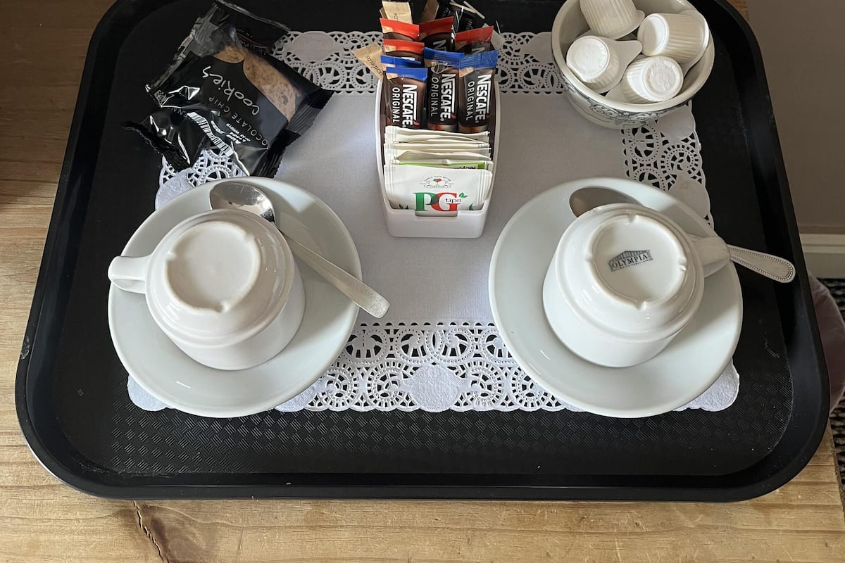A tray with a box of Nescafe Original coffee, a pack of PG tea, and a bowl of white ceramic cups on a table with a lace doily.