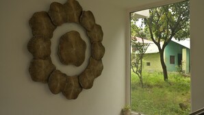 Un adorno mural con diseño circular, una ventana con vistas a zonas verdes y un edificio, y un estante con objetos.
