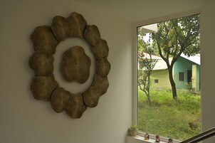 Un adorno mural con diseño circular, una ventana con vistas a zonas verdes y un edificio, y un estante con objetos.