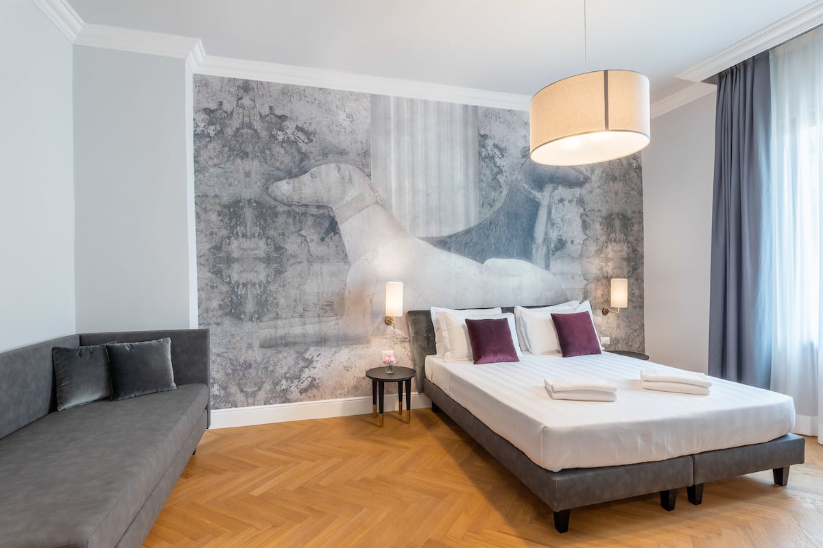 Un dormitorio moderno con una cama grande, un sofá gris, una mesita redonda y un mural en la pared.