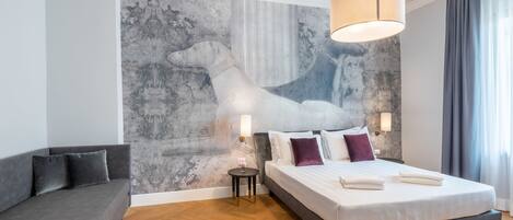 A modern bedroom with a large bed, a grey sofa, a small round table, and a wall mural.
