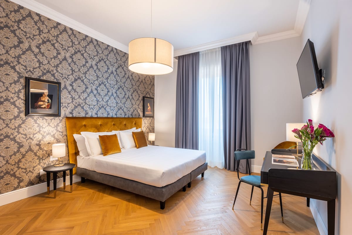 Habitación de hotel con una cama grande, un escritorio con silla, un televisor montado en la pared y una ventana con cortinas.