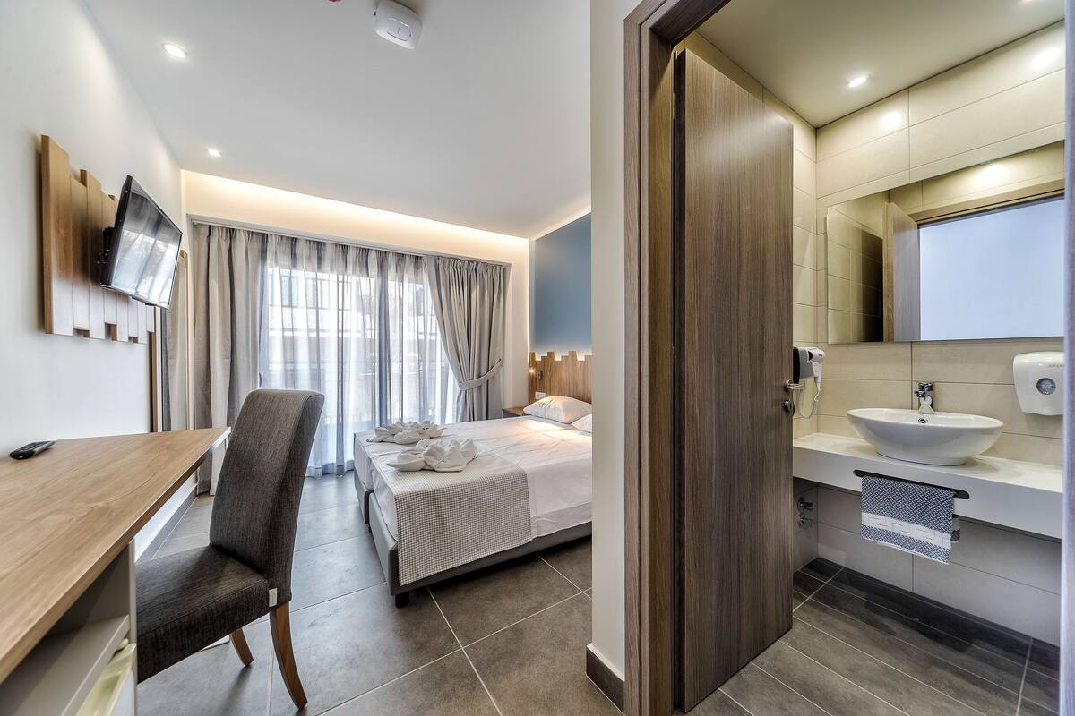 A hotel room with a bed, a desk, a chair, a TV mounted on the wall, a bathroom with a sink and mirror, and a window with curtains.