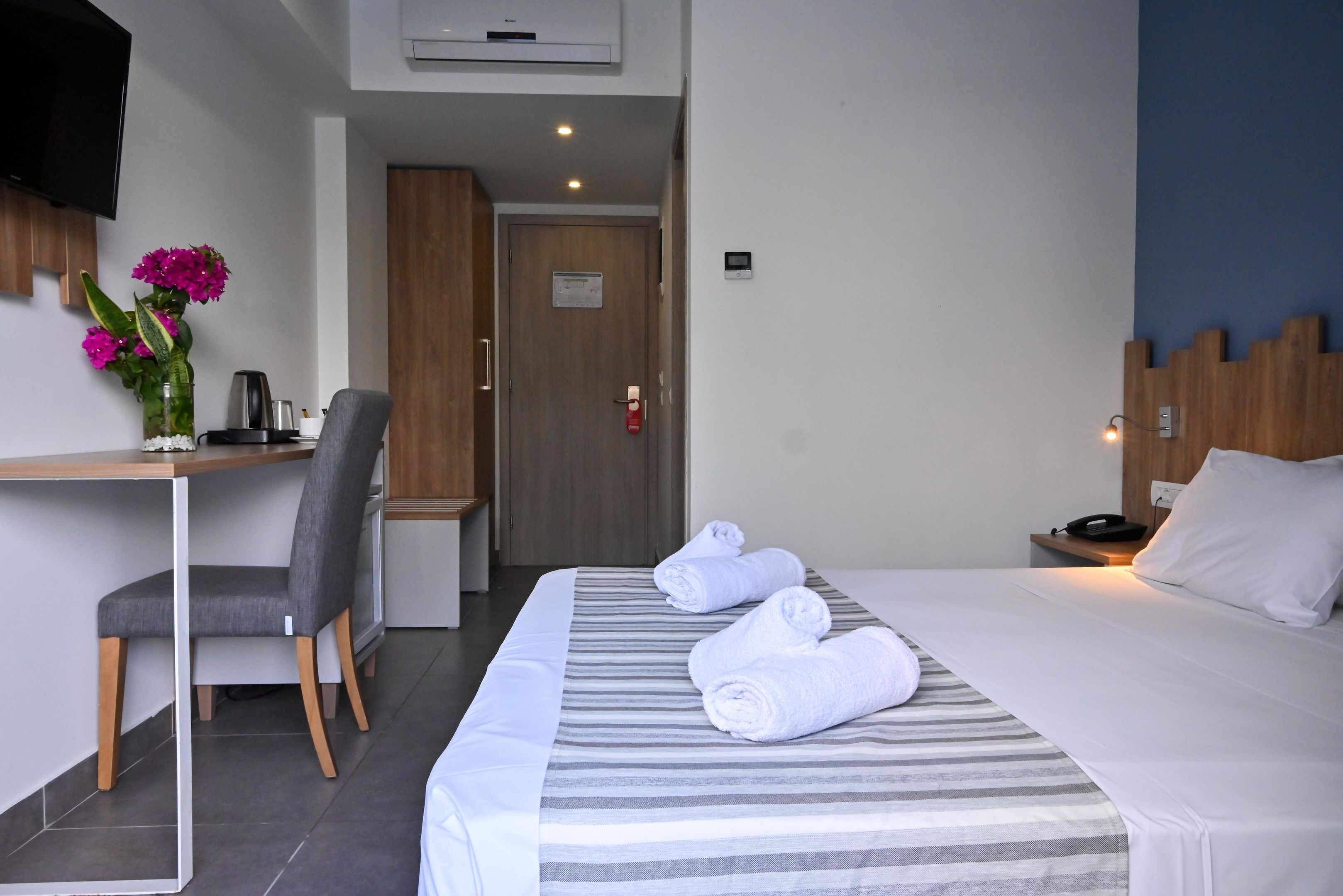A hotel room with a bed, a desk with a chair, a TV mounted on the wall, and a door with a fire alarm.