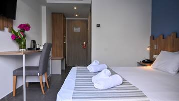 A hotel room with a bed, a desk with a chair, a TV mounted on the wall, and a door with a fire alarm.