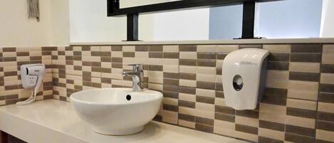 Une salle de bain avec un lavabo blanc, un miroir, un distributeur de savon et un sèche-cheveux fixé au mur.